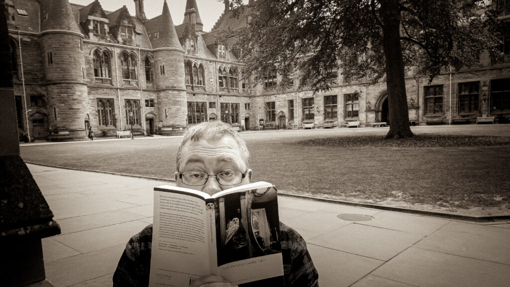 Image of the author, face obscured reading a book of victorian ghost stories, in front of a backdrop of neo-gothic architecture