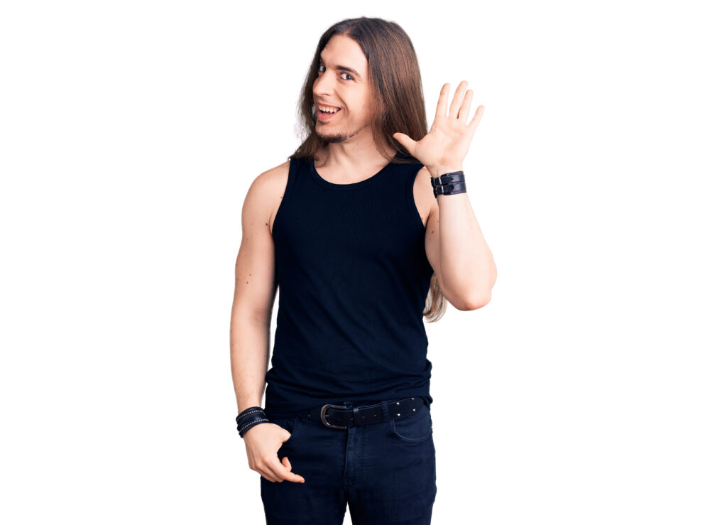 Young adult man with long hair wearing goth style with black clothes waiving saying hello happy and smiling, friendly welcome gesture